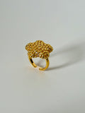 A gold-colored adjustable ring with a textured, woven floral or cloud-like top, photographed on a neutral light gray background.