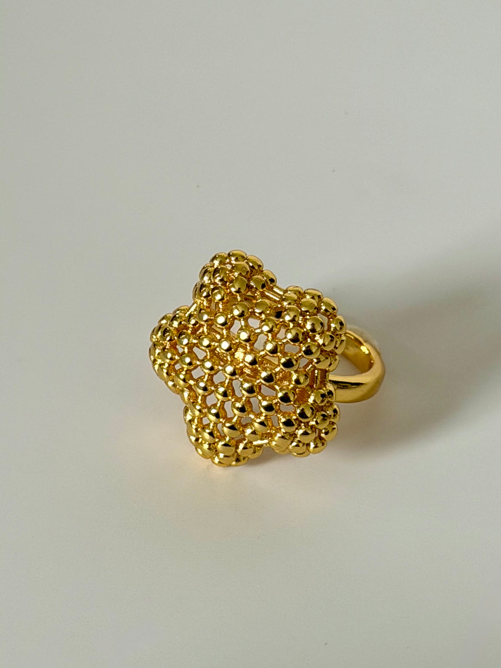 A textured gold ring featuring a five-petal flower design with an intricate openwork mesh pattern and a smooth gold band, shown on a neutral light gray background.