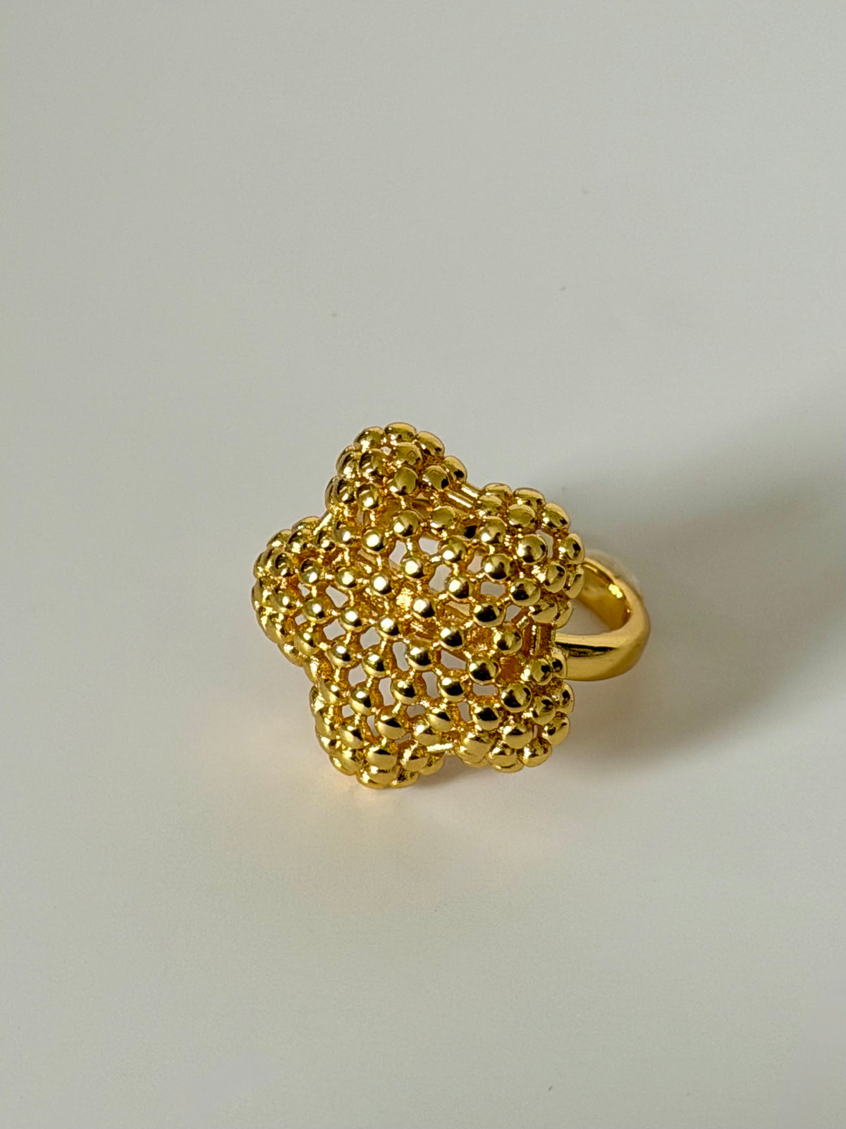 A textured gold ring featuring a five-petal flower design with an intricate openwork mesh pattern and a smooth gold band, shown on a neutral light gray background.