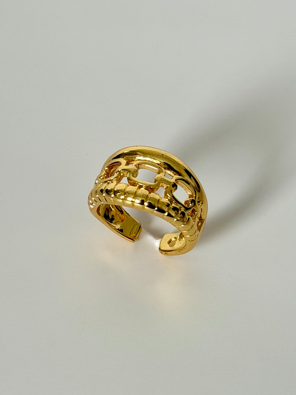 A polished gold open cuff ring with a textured, interlocking link design, photographed on a neutral light gray background.