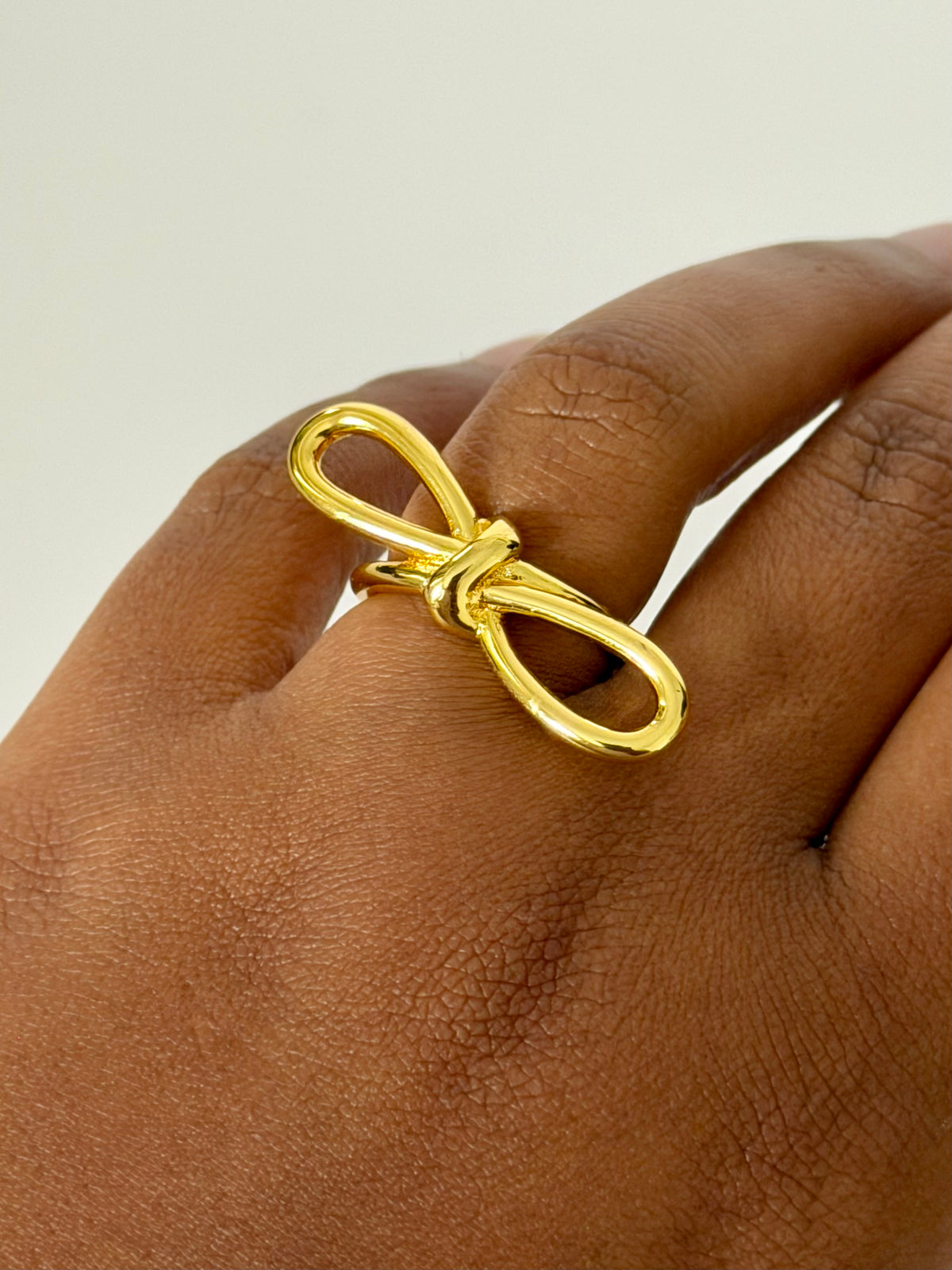 A polished gold ring shaped like a stylized bow or infinity knot, shown worn on a person's finger against a plain light background.