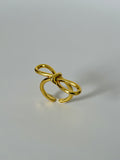 A gold-colored metal ring with a sculptural, open bow-like design, photographed on a neutral light gray background.
